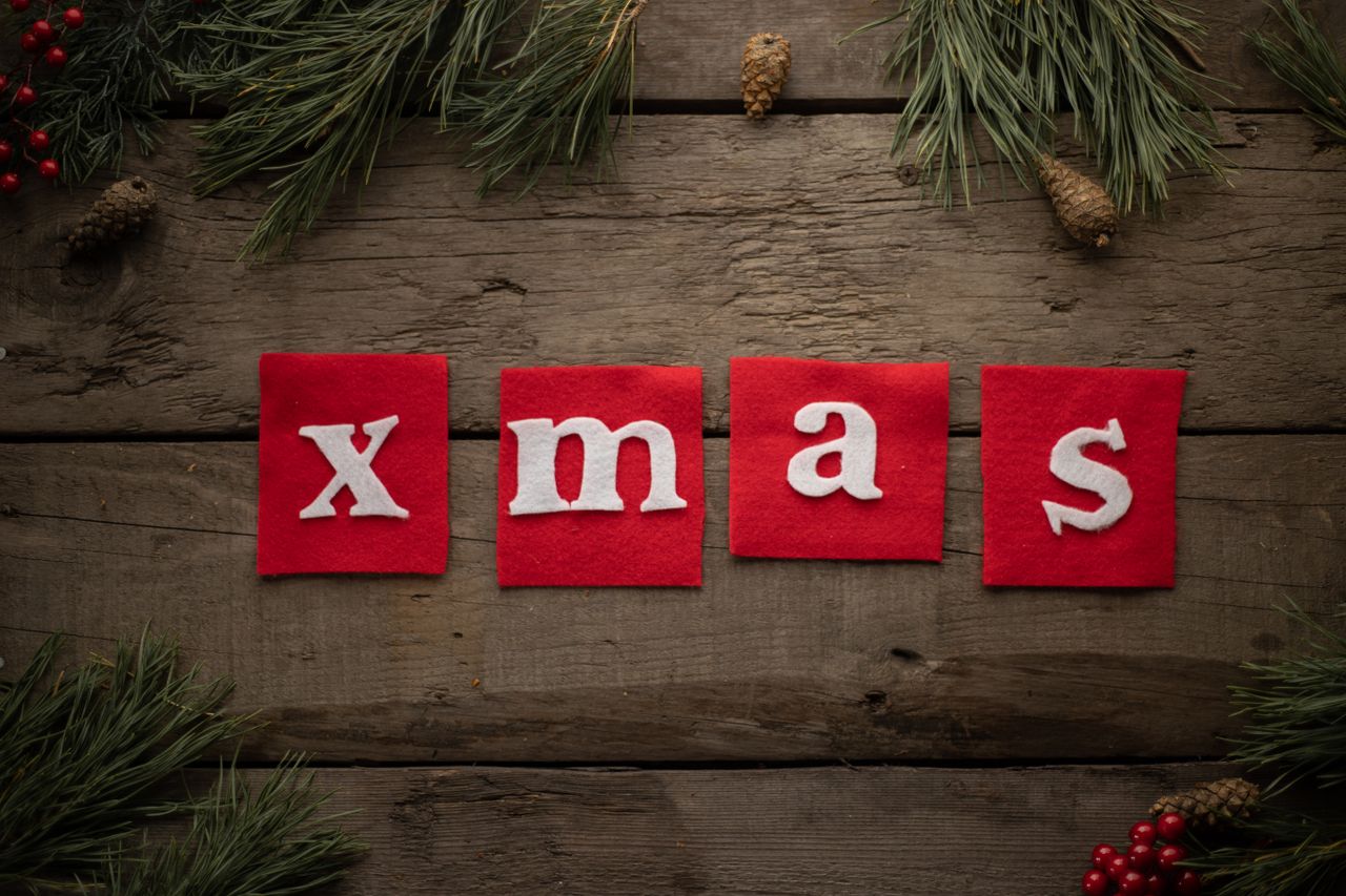Top view arrangement of white Xmas inscription on red square fabric placed on plank table  surrounded by coniferous tree branches and cones during festive season